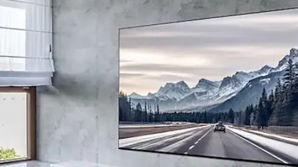 Pokój z oknem z żaluzjami i wiszącym telewizorem, na którym wyświetlany jest krajobraz górski z drogą i lasem.