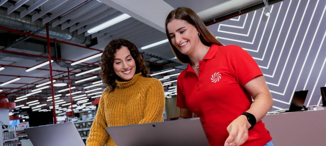 Dwie kobiety patrzą na laptopa w sklepie. Jedna w żółtym swetrze, druga w czerwonej koszulce polo z logo. W tle półki i oświetlenie.