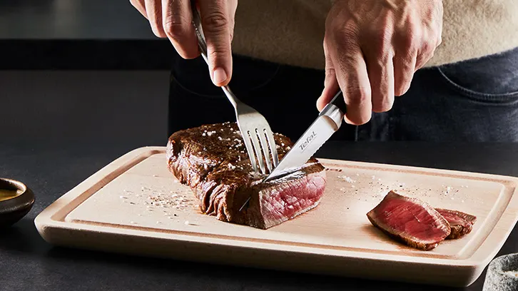 Osoba kroi stek nożem i widelcem na desce do krojenia. Obok widać małą miseczkę i przyprawy.