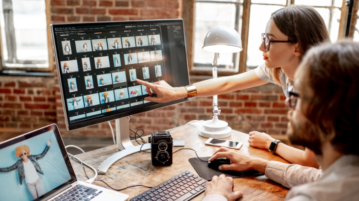 Dwie osoby przy biurku z komputerami. Na ekranie monitora widoczne zdjęcia, kobieta pokazuje palcem na ekran. Obok lampa i aparat.