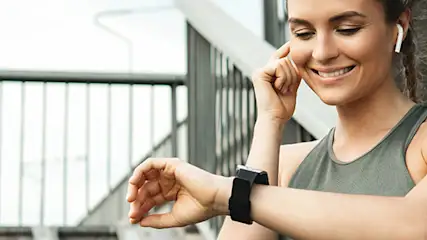 Uśmiechnięta kobieta w sportowym topie patrzy na czarny smartwatch na tle betonowych schodów z metalową barierką.