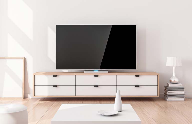 Pokój z czarnym telewizorem na białej komodzie z sześcioma szufladami. Obok lampa na książkach i biały stolik z wazonem.