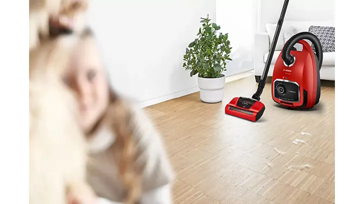 Na drewnianej podłodze stoi czerwony odkurzacz, obok doniczka z zieloną rośliną, w tle biała ściana i sofa.