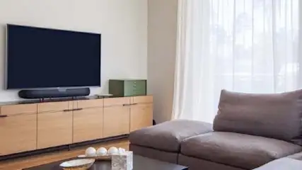 Salon z brązową kanapą, szafką pod telewizorem i zasłonami w oknie. Na szafce stoi telewizor i głośnik.