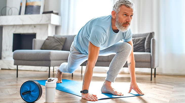 Dojrzały wysportowany mężczyzna w treningowym stroju i smartwatchem, wykonuje wykrok na macie do jogi w salonie.