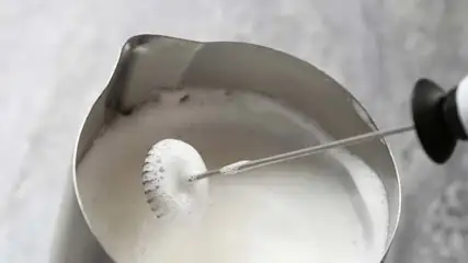 Pienik w metalowym dzbanku. W środku biała ciecz i spieniacz do mleka. Tło jest szare.