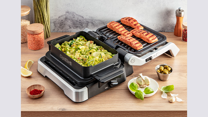 Na drewnianym stole stoi grill elektryczny z potrawami: cukinią i grillowanym łososiem. Obok są przyprawy, czosnek, oliwki i cytryna.