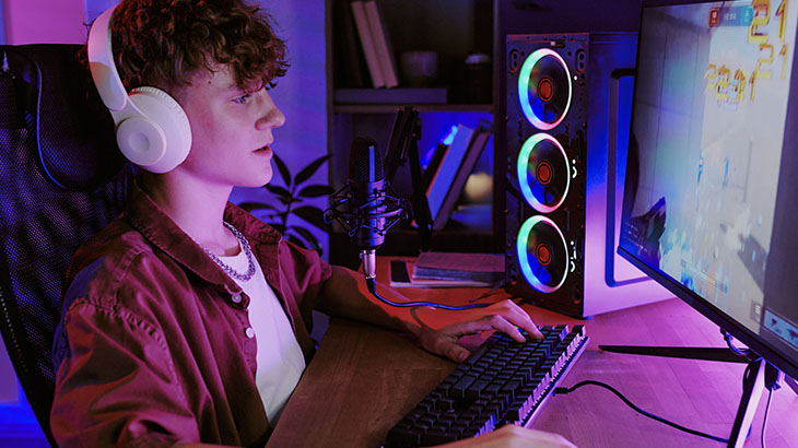 Osoba gra przy komputerze, ma na uszach słuchawki, w tle komputer, monitor i klawiatura.