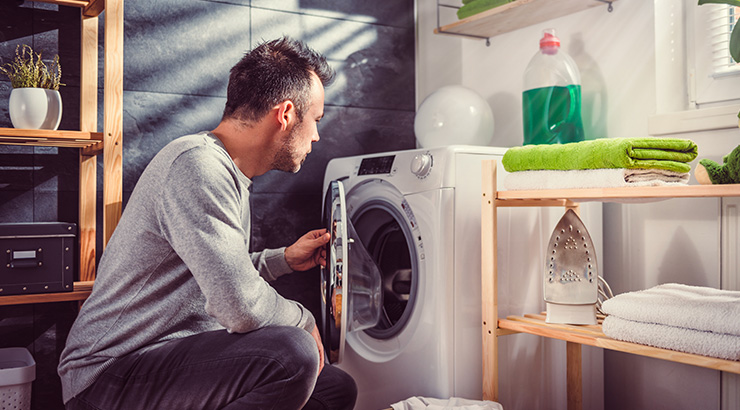 Mężczyzna przy otwartej pralce w łazience. W tle półki z ręcznikami, żelazkiem i płynem do prania.