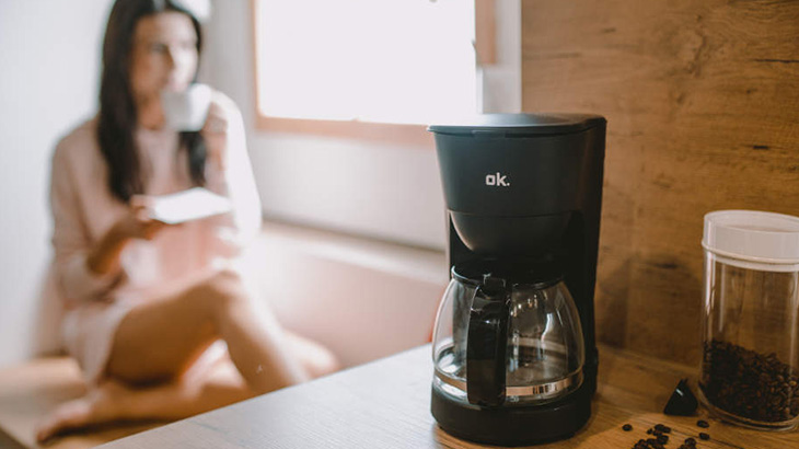 Czarny ekspres do kawy obok pojemnika z ziarnami kawy na blacie. Kobieta pije kawę w tle, siedząc przy oknie.