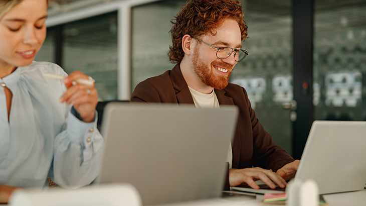 Rudowłosy mężczyzna siedzi obok kobiety, uśmiechają się i pracują przy laptopach