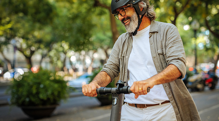 Mężczyzna w kasku i okularach jedzie hulajnogą po ulicy, w tle zieleń drzew i pojazdy.