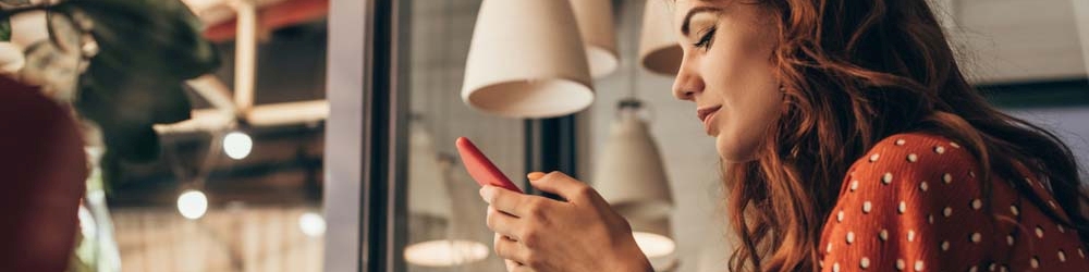 Kobieta w czerwonej bluzce w kropki używa telefonu. W tle lampy i okno z odbiciami.