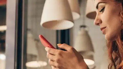 Kobieta w czerwonej bluzce w kropki używa telefonu. W tle lampy i okno z odbiciami.