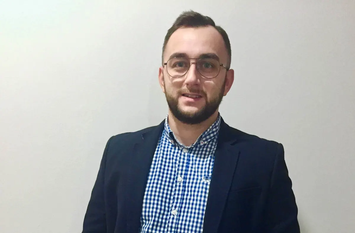 Mężczyzna w okularach i marynarce na tle jasnej ściany. Ma na sobie koszulę w niebiesko-białą kratę.