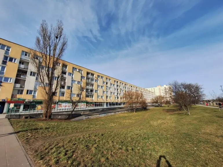 Długi, żółty blok mieszkalny z oknami, drzewa bez liści, zielony trawnik i niebieskie niebo z chmurami.