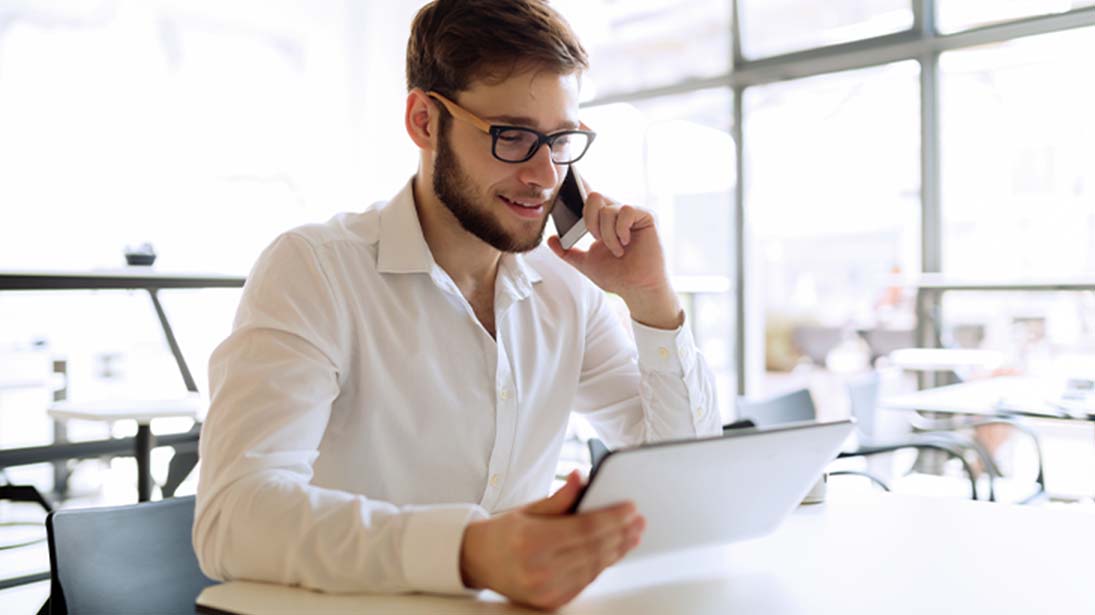 Mężczyzna rozmawia przez telefon, trzymając w dłoni tablet, siedząc przy biurku.