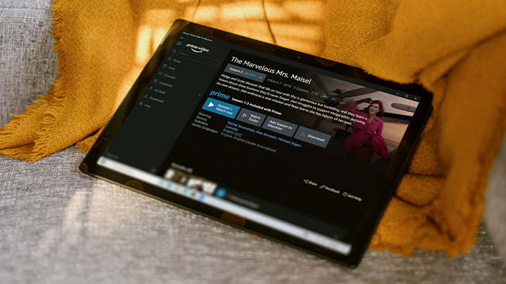 Czarny tablet leży na szarej sofie obok żółtej tkaniny. Na ekranie tabletu widoczna jest aplikacja Amazon Prime Video.