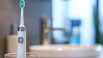 Elektryczna szczoteczka do zębów w szklance. Tło: umywalka i bateria.