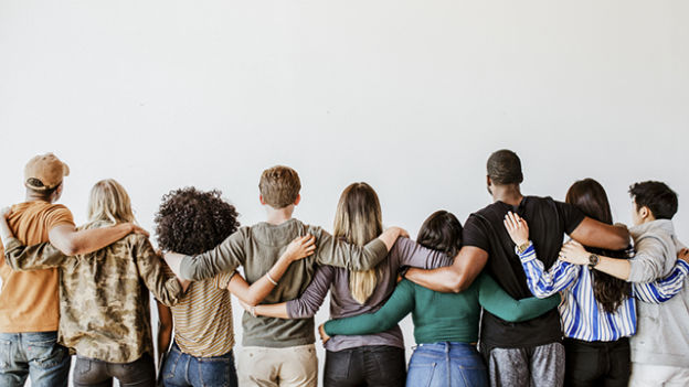 Dziewięć osób stoi ramię w ramię, tyłem do widza, na tle białej ściany. Noszą różne ubrania, od koszulek po bluzy i dżinsy.