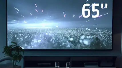 Salon z dużym telewizorem 65 cali, na którym wyświetlane są efekty świetlne. Widoczne osoby oglądające, lampa i regał.