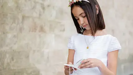 Dziewczynka w białej sukience trzyma różaniec na tle ściany z cegieł. Na głowie ma wianek z kwiatów.