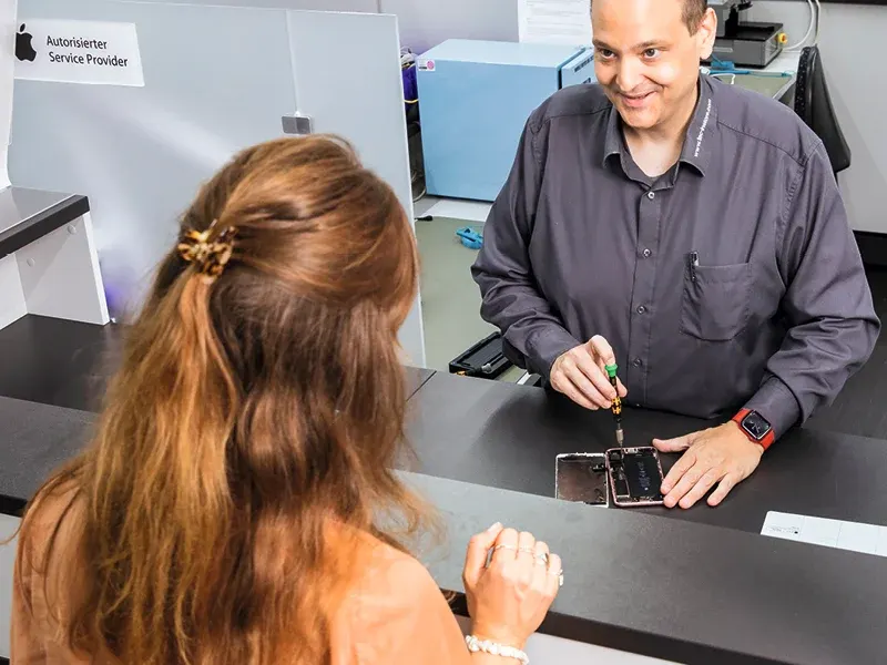 Kobieta stoi przy ladzie, obserwując mężczyznę w szarej koszuli, który naprawia telefon śrubokrętem na czarnym blacie.