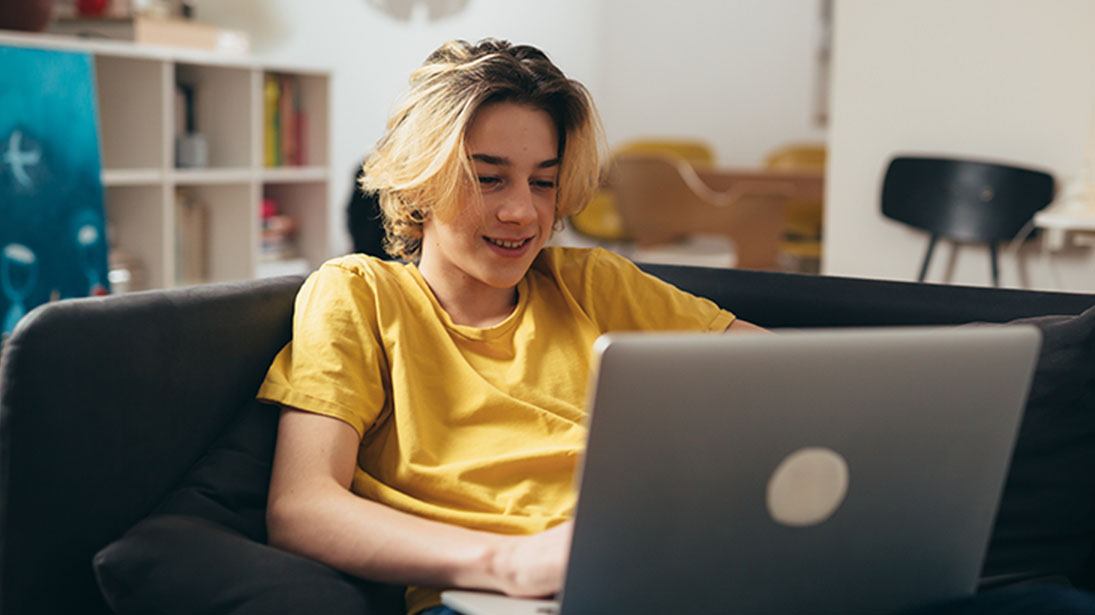 Chłopiec w żółtej koszulce siedzi na szarej kanapie, używając laptopa. W tle widać regał i krzesło.