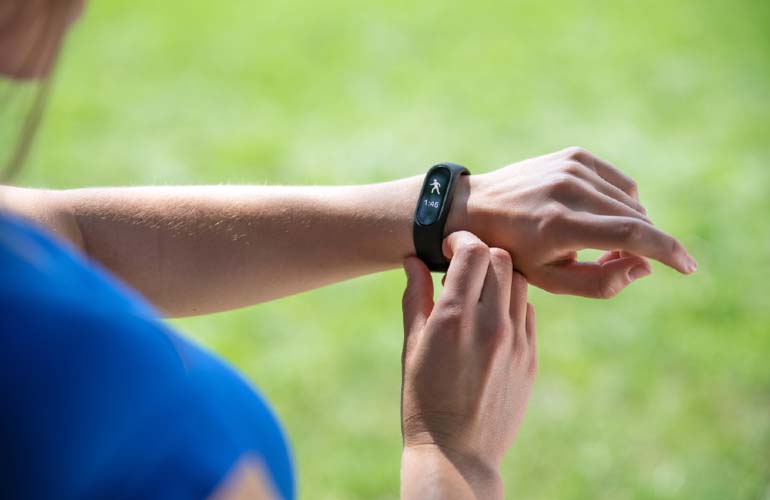 Osoba w niebieskiej koszulce sprawdza czarny smartwatch na nadgarstku, na tle zieleni.