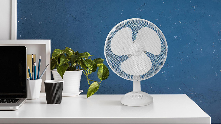 Biurko z laptopem, wentylatorem, rośliną doniczkową, kubkiem i przyborami do pisania na tle niebieskiej ściany.