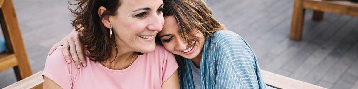Kobieta w różowej koszulce obejmuje dziewczynę w niebieskiej koszuli w paski. Uśmiechają się siedząc na drewnianej ławce.