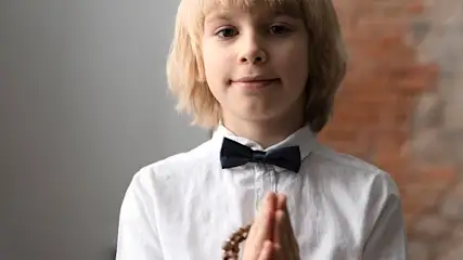 Chłopiec w białej koszuli i czarnej muszce składa dłonie. W dłoniach trzyma różaniec. W tle szara ściana i cegła.