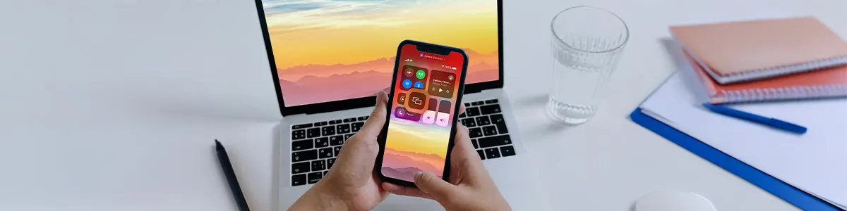 Smartfon z menu ustawień iPhone'a na tle MacBooka, jakichś zeszytów i szklanki z wodą