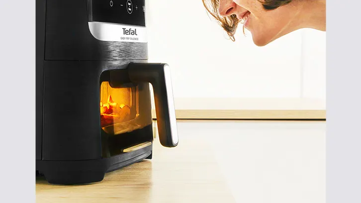 Czarna frytownica Tefal z nagrzanym wnętrzem stoi na blacie. Nad nią pochyla się osoba. Tło jest jasne.