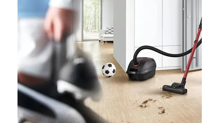 Odkurzacz w akcji na drewnianej podłodze obok czarno-białej piłki. W tle jasny pokój z białymi meblami i oknem.