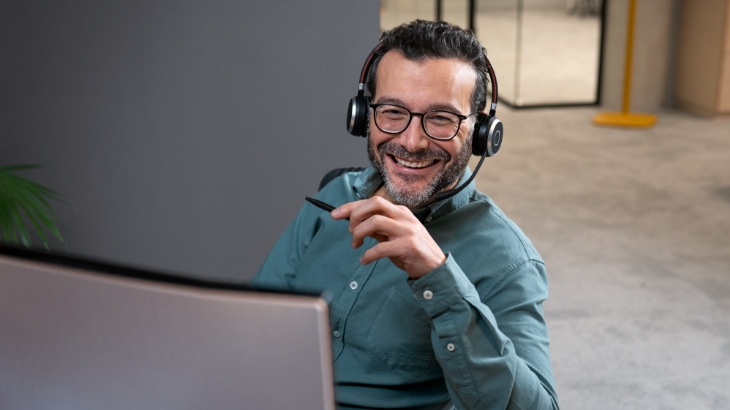 Mężczyzna w okularach i słuchawkach uśmiecha się, trzymając długopis. Przed nim laptop, w tle biuro z szarą ścianą.