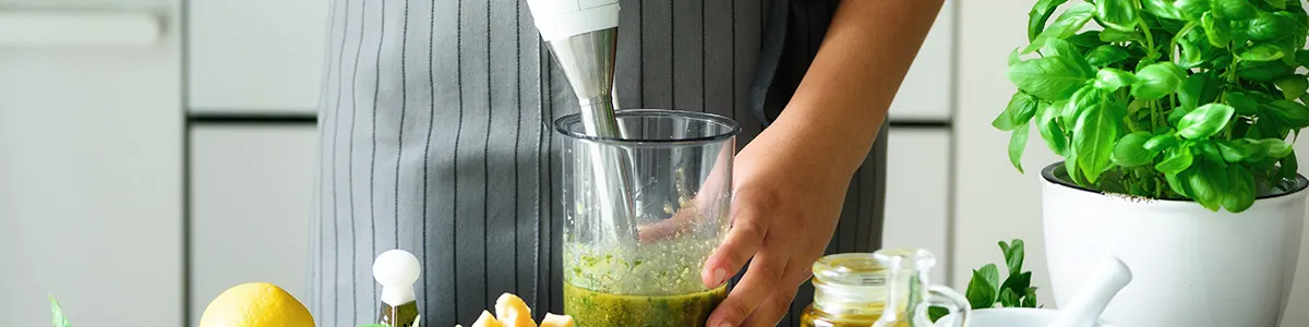 Osoba w fartuchu używa blendera w wysokiej szklance z zieloną cieczą, obok cytryna i bazylia w białej doniczce.