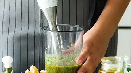 Osoba w fartuchu używa blendera w wysokiej szklance z zieloną cieczą, obok cytryna i bazylia w białej doniczce.