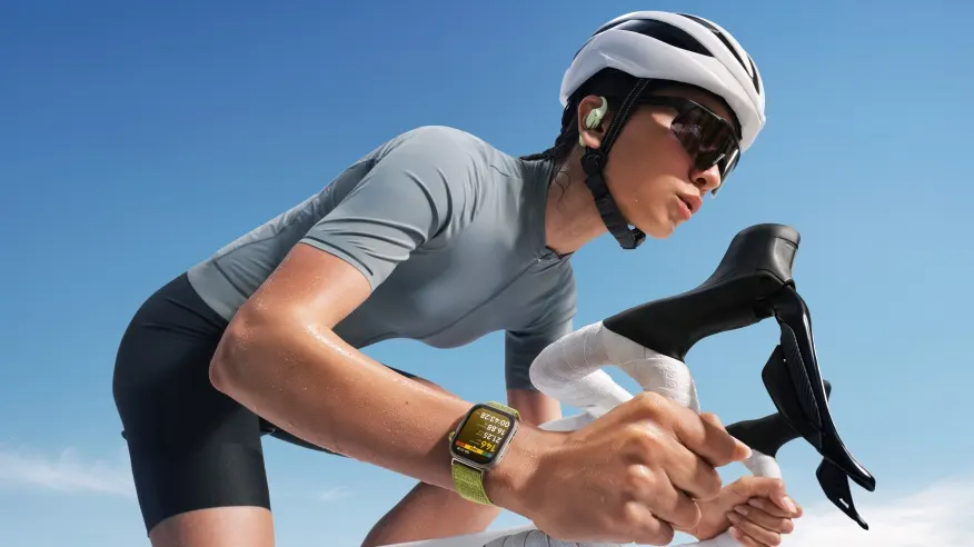 Kobieta na rowerze w kasku i okularach przeciwsłonecznych. Nosi szarą koszulkę, czarne spodenki i zegarek na zielonym pasku.