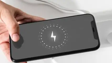 Dłoń trzyma czarny smartfon z symbolem ładowania na ekranie, leżący na białej ładowarce indukcyjnej na białym tle.