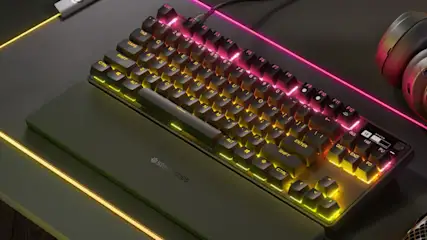 Czarna klawiatura na biurku, podświetlona na różowo i żółto, obok słuchawki i podkładka z logo SteelSeries.