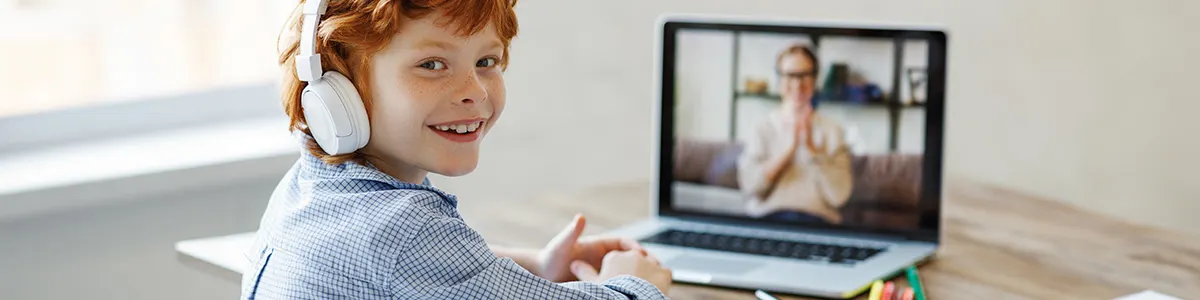 Rudowłosy chłopiec siedzi w białych słuchawkach i rozmawia z kimś przez videocalla na laptopie