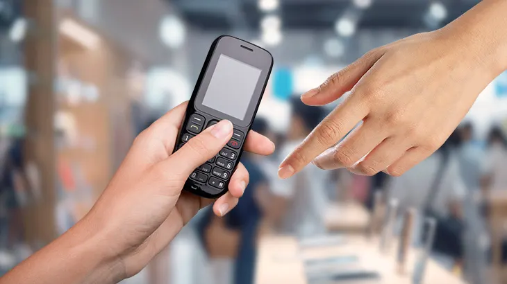 Dłoń trzyma czarny telefon z klawiaturą. Druga dłoń wyciąga palec wskazujący w kierunku telefonu. Tło jest rozmyte.