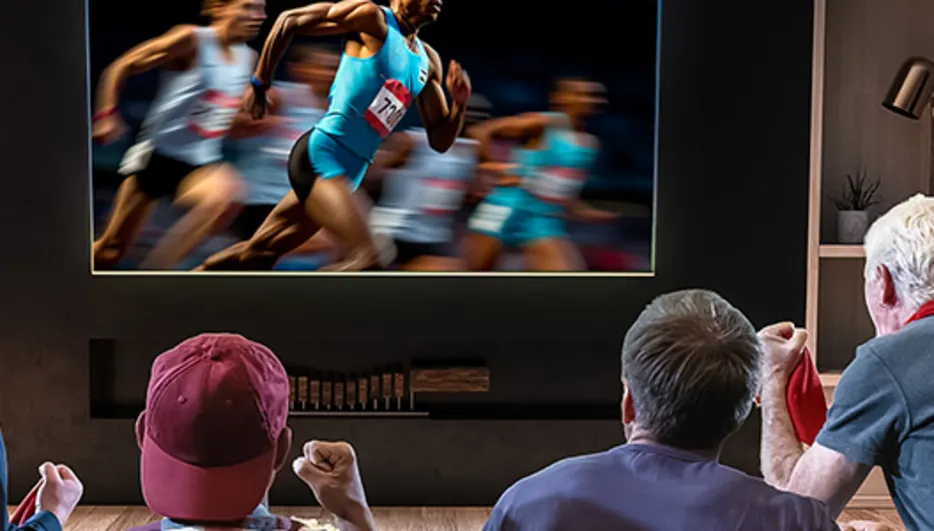 Czterech mężczyzn ogląda biegaczy na telewizorze w pokoju. Półki z książkami i dekoracjami stoją za nimi.