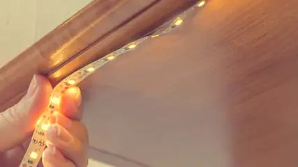 Dłoń trzyma taśmę LED z ciepłym światłem, przyklejając ją pod drewnianą półką.