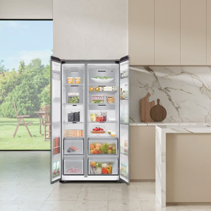 Otwarta, dwudrzwiowa lodówka stoi w jasnej kuchni obok okna z widokiem na zieleń. Wnętrze wypełnione produktami spożywczymi.