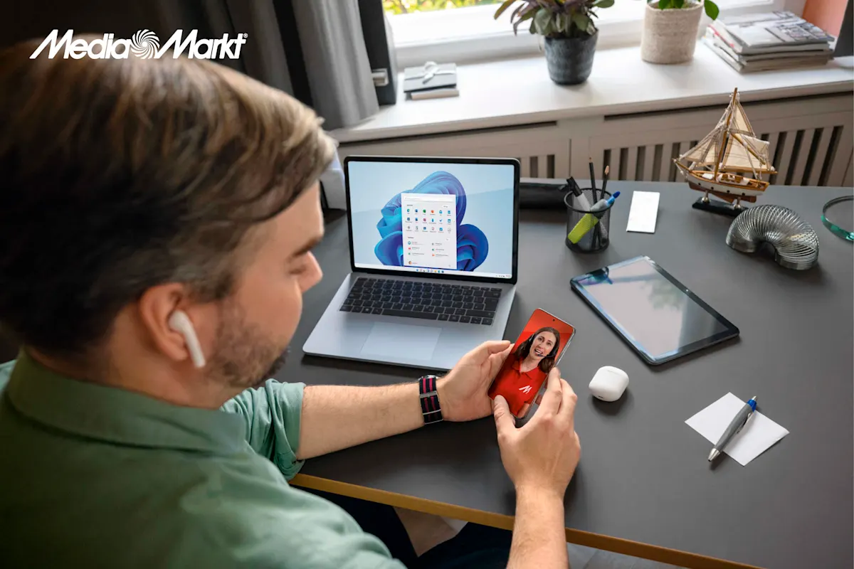 Mężczyzna w słuchawce rozmawia przez telefon. Na biurku leżą: laptop, tablet, słuchawki, długopis i model żaglówki.
