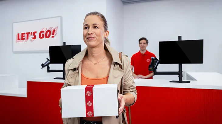 Kobieta z paczką w ręku. W tle kasy sklepowe, sprzedawca w czerwonej koszulce. Na ścianie napis "Let's go!"