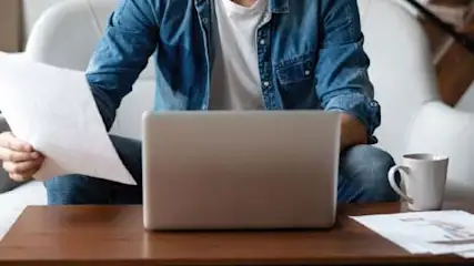 Osoba w dżinsowej koszuli siedzi z laptopem i papierami na stole, obok kubek i roślina doniczkowa.