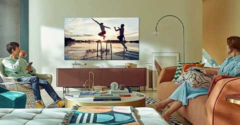 Salon z telewizorem pokazującym osoby skaczące do wody, kanapa z dwiema osobami oglądającymi, stolik i lampa łukowa.